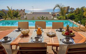 Un área de comedor al aire libre con vista a la piscina, mesas y sillas de madera, y una exhibición de frutas tropicales.