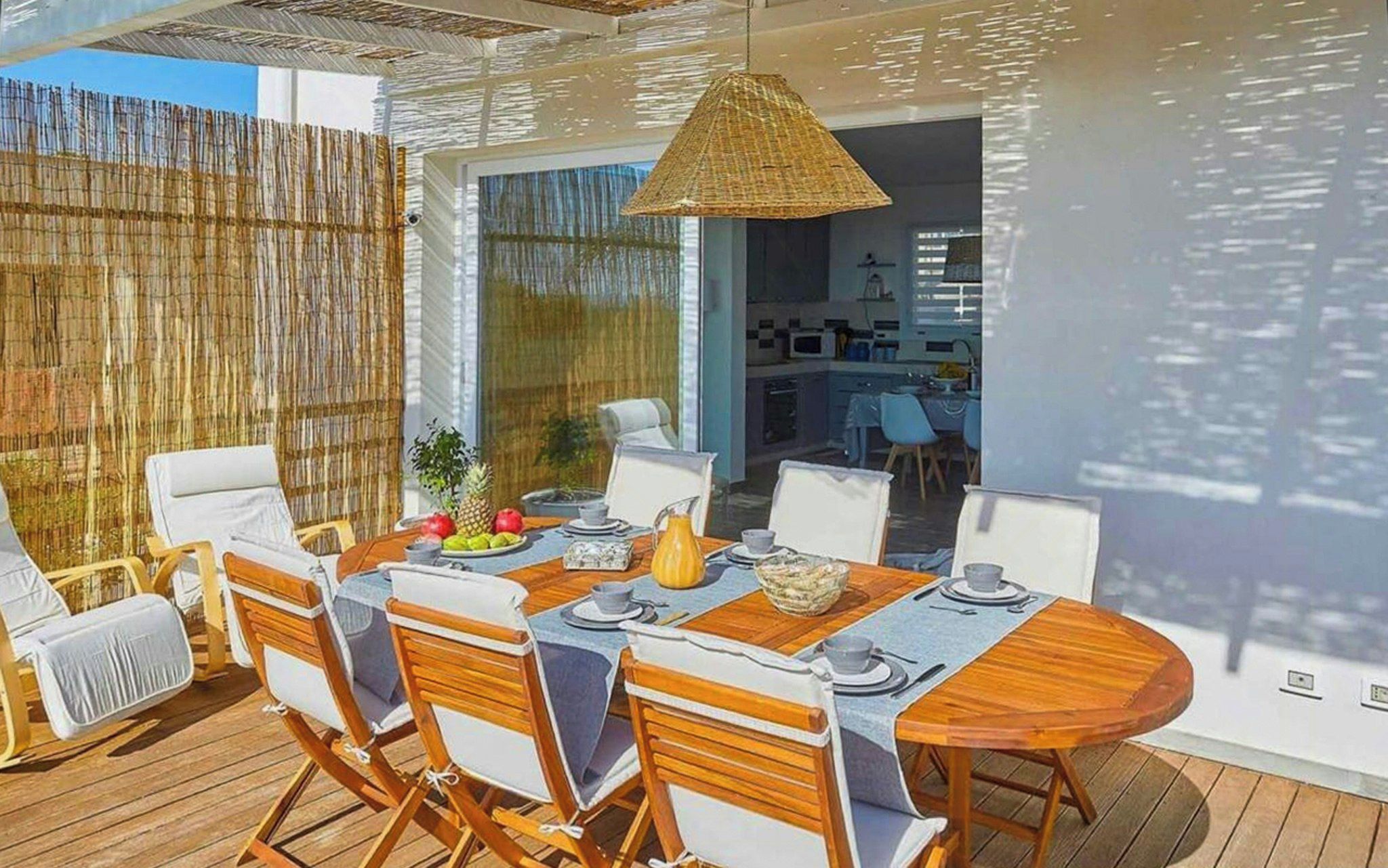 An outdoor dining area with a wooden table and chairs, a woven pendant light, and a view into the adjacent kitchen.