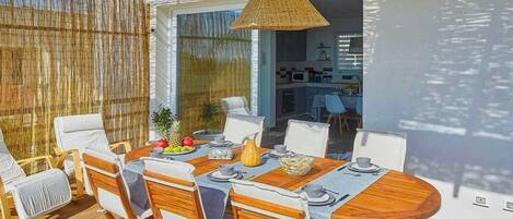 An outdoor dining area with a wooden table and chairs, a woven pendant light, and a view into the adjacent kitchen.