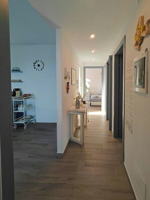 Un pasillo con suelo de madera, un reloj de pared blanco, una pequeña mesa con un jarrón, y una cama a lo lejos.