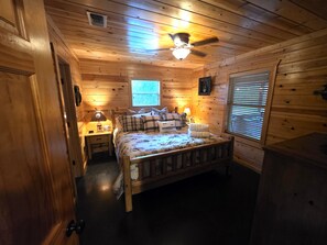 Quarto com painéis de madeira, uma cama, mesa de cabeceira, abajur e ventilador de teto.