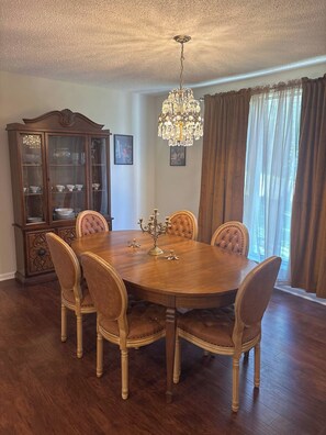 Ein Esszimmer mit Holztisch, sechs Polsterstühlen, einer Kristallleuchter, einer Speisekammer und einem Fenster mit Vorhängen.