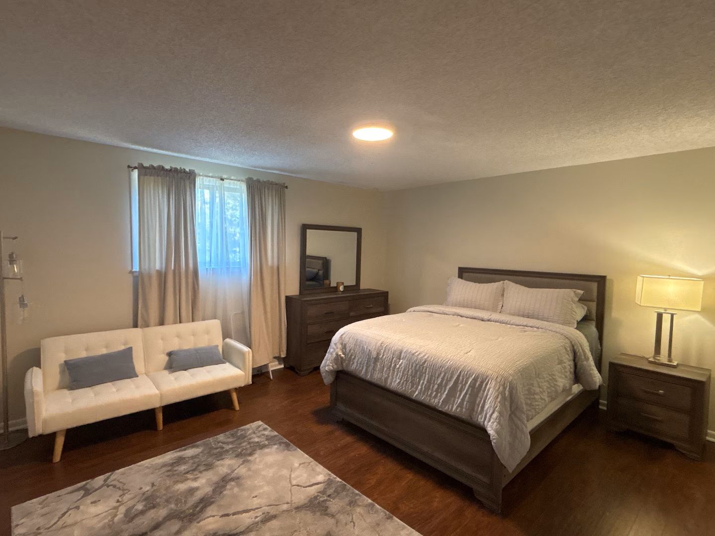 A bedroom with a bed, a nightstand, a dresser, a sofa, and a window with curtains.