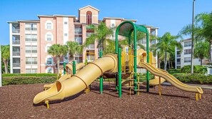 Un parque infantil con toboganes y estructuras para escalar, rodeado de un área ajardinada con árboles y un edificio al fondo.