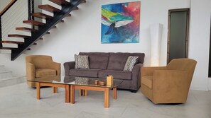 A living room with a sofa, two armchairs, a glass coffee table, and a colorful abstract painting.