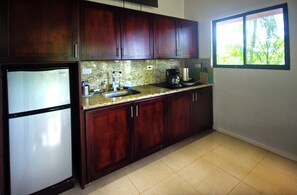 A kitchen with dark wooden cabinets, a granite countertop, a refrigerator, and a window with a view of greenery.