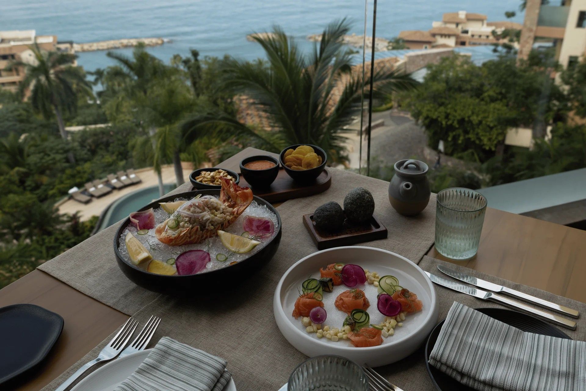 Une table à manger dressée avec des plats de fruits de mer, une théière, et une vue sur la mer et les bâtiments.