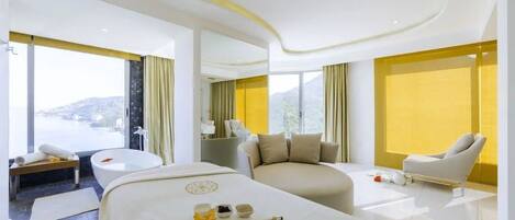 A modern hotel room with a large bed, a sitting area with a sofa, a bathtub, and a view of the outdoors.