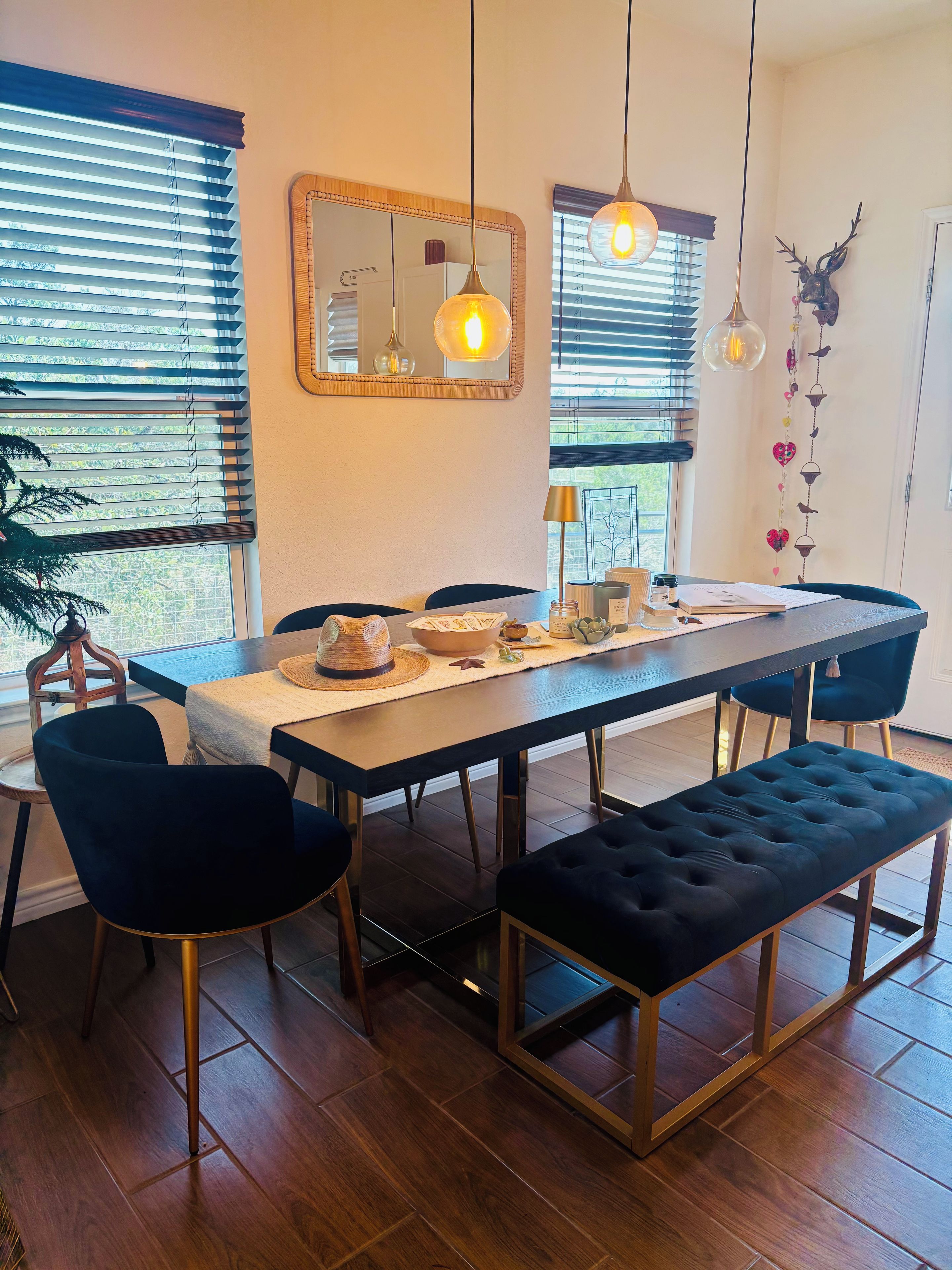 A dining area with a long table, chairs, and benches. There are hanging lights, a mirror, and a wall decoration.
