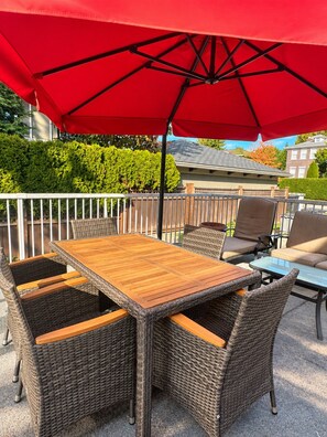 An outdoor patio with a wooden table and wicker chairs, a red umbrella, and a white railing. - Grand King Suite | 5-10 Min to Airport & Downtown (Vancouver)