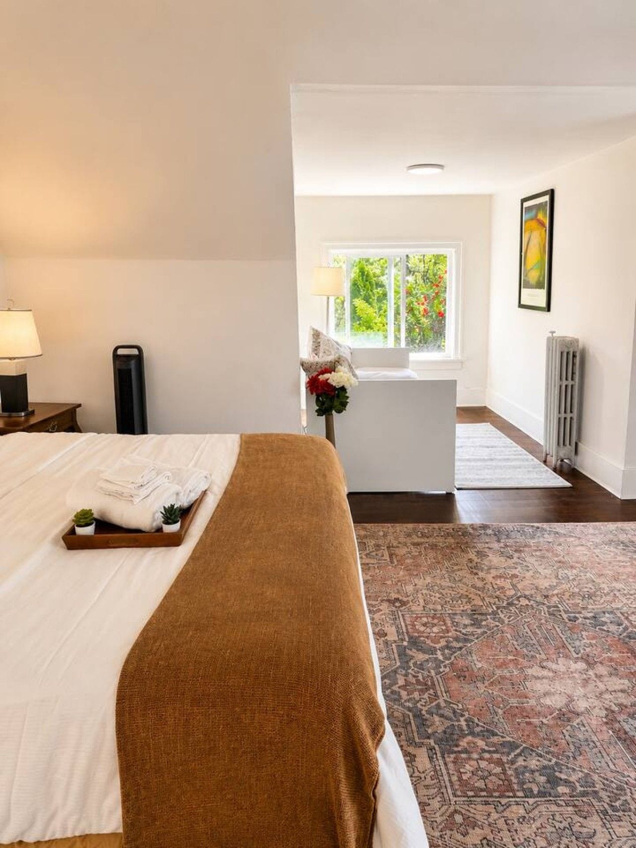 Une chambre d’hôtel avec un lit, une table de chevet avec une lampe, un radiateur, une fenêtre donnant sur un espace verdoyant et un tapis.