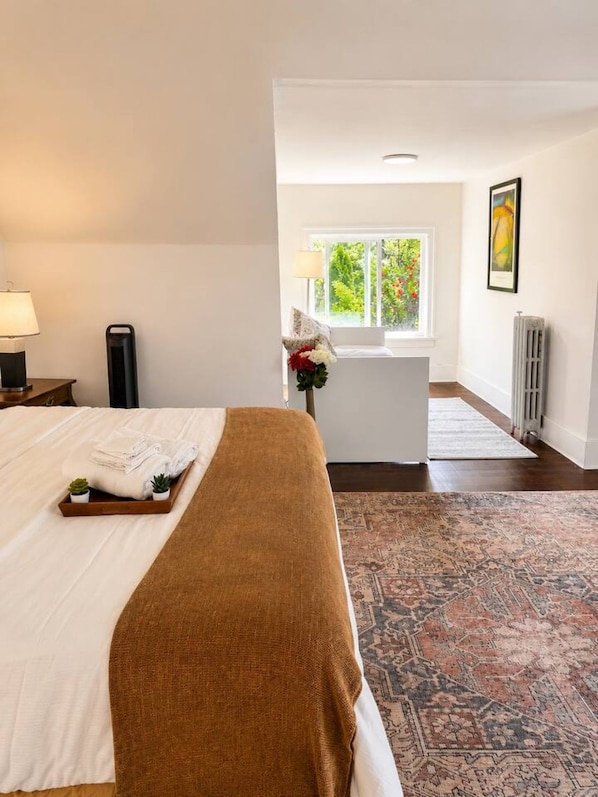 A hotel room with a bed, a bedside table with a lamp, a radiator, a window with a view of greenery, and a rug.