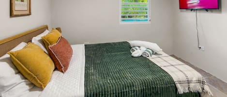 A hotel room with a bed, pillows, a green blanket, a TV, and a window with blinds.