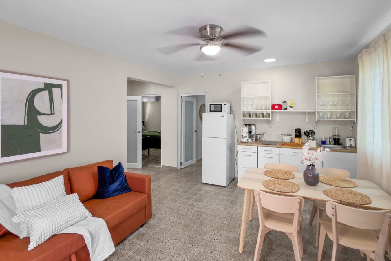 Een knus woongedeelte met een kitchenette, een eethoek en een zithoek met een bank en een plafondventilator.