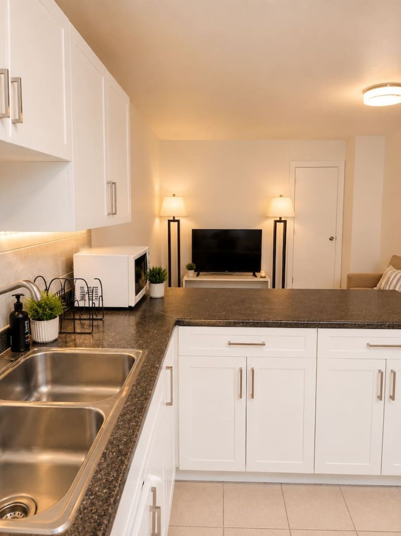 A modern kitchen with white cabinetry, a stainless steel sink, a microwave, and two floor lamps.
