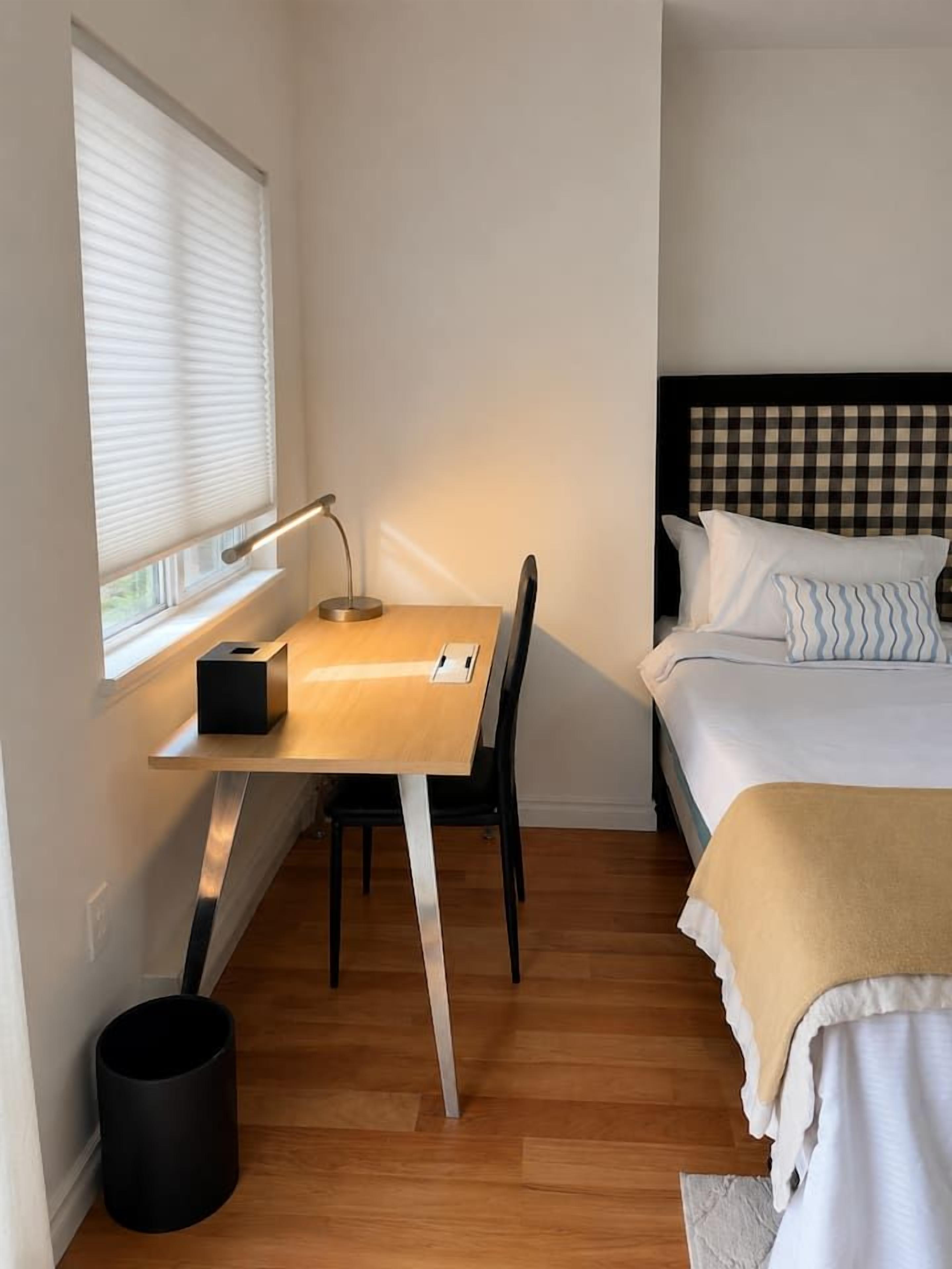 A compact room with a wooden desk, a chair, a bed with a checkered headboard, and a window with blinds.