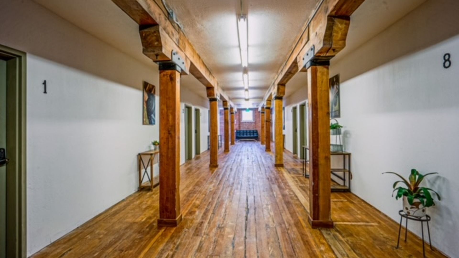 Un pasillo con suelo de madera, vigas a la vista y puertas numeradas a la izquierda.