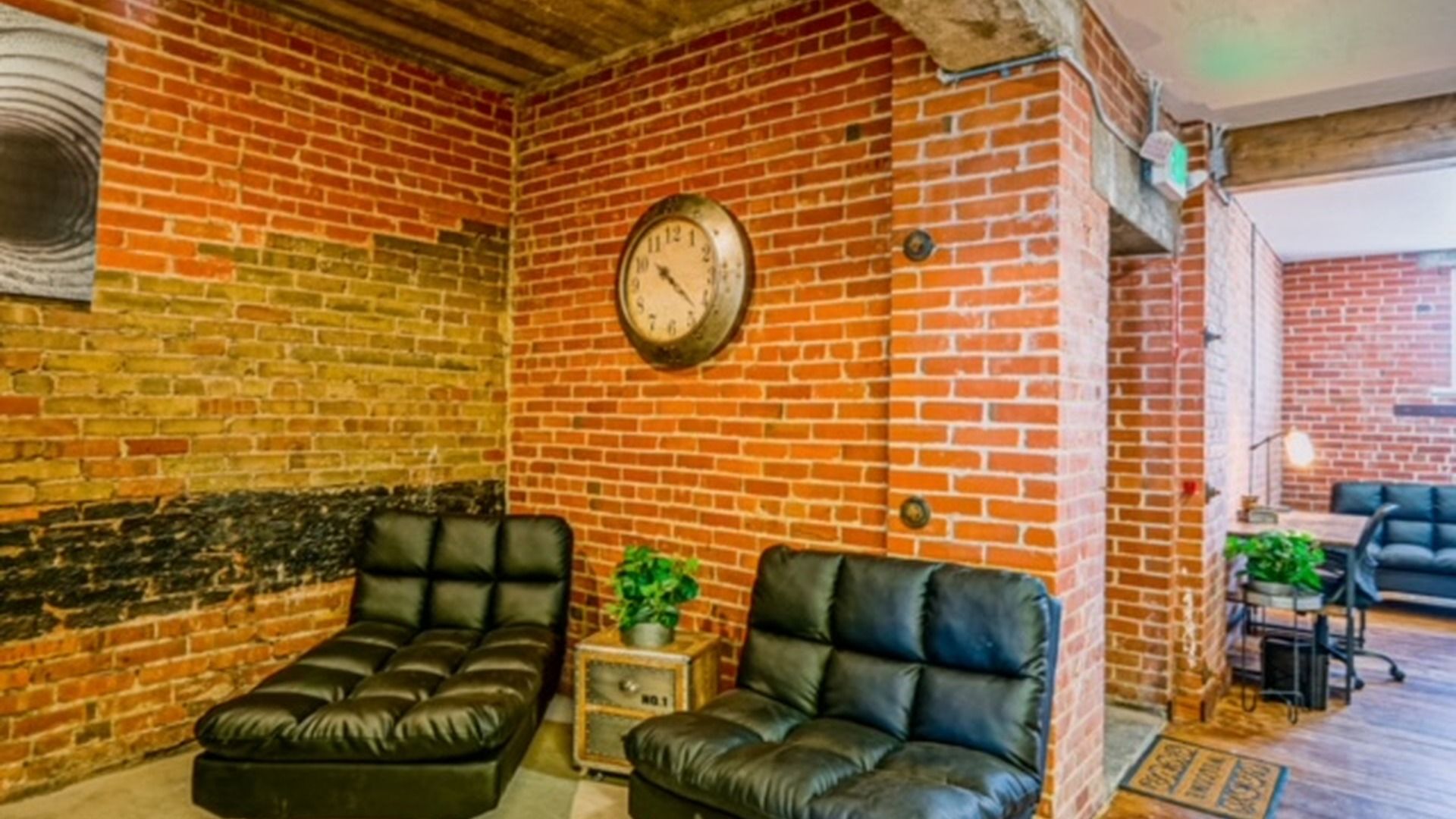 Una habitación con paredes de ladrillo, un reloj redondo, dos sofás de cuero y una pequeña mesa con una planta.