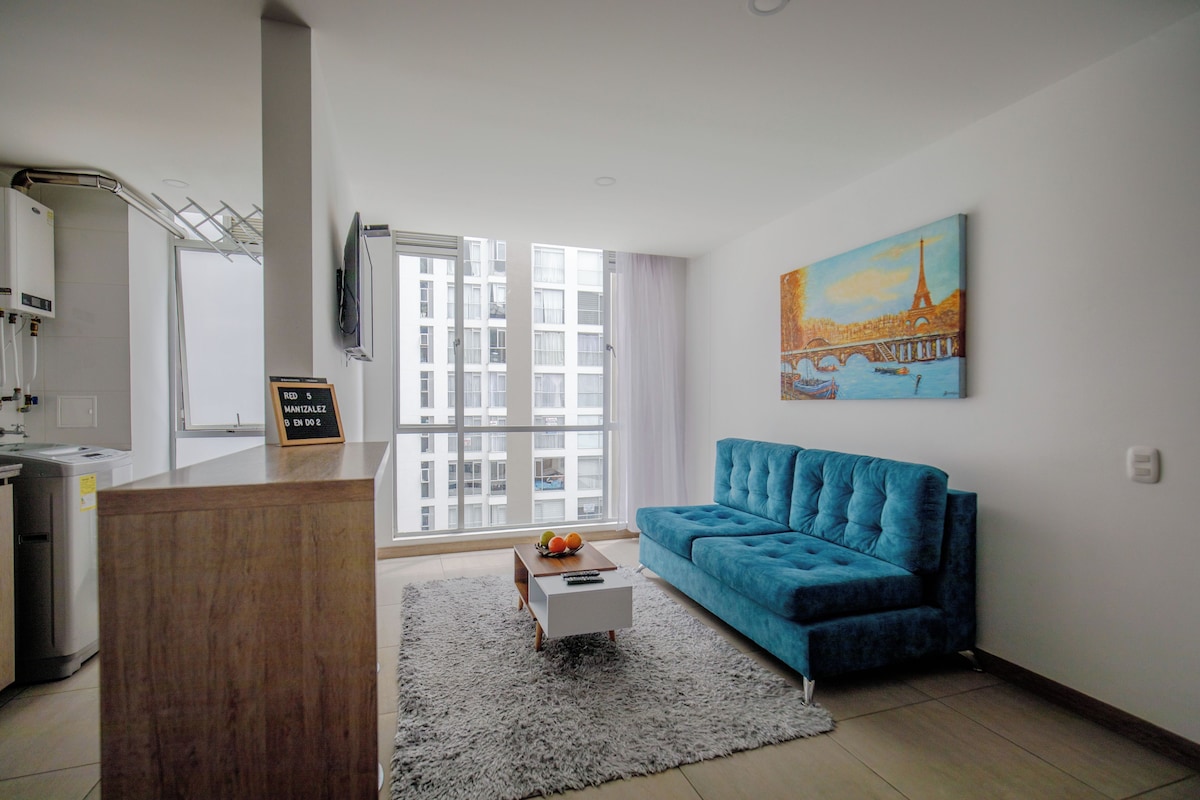 Una sala moderna con un sofá azul, una mesa de centro con frutas, un cuadro de un paisaje urbano y vistas de edificios a través del ventanal.