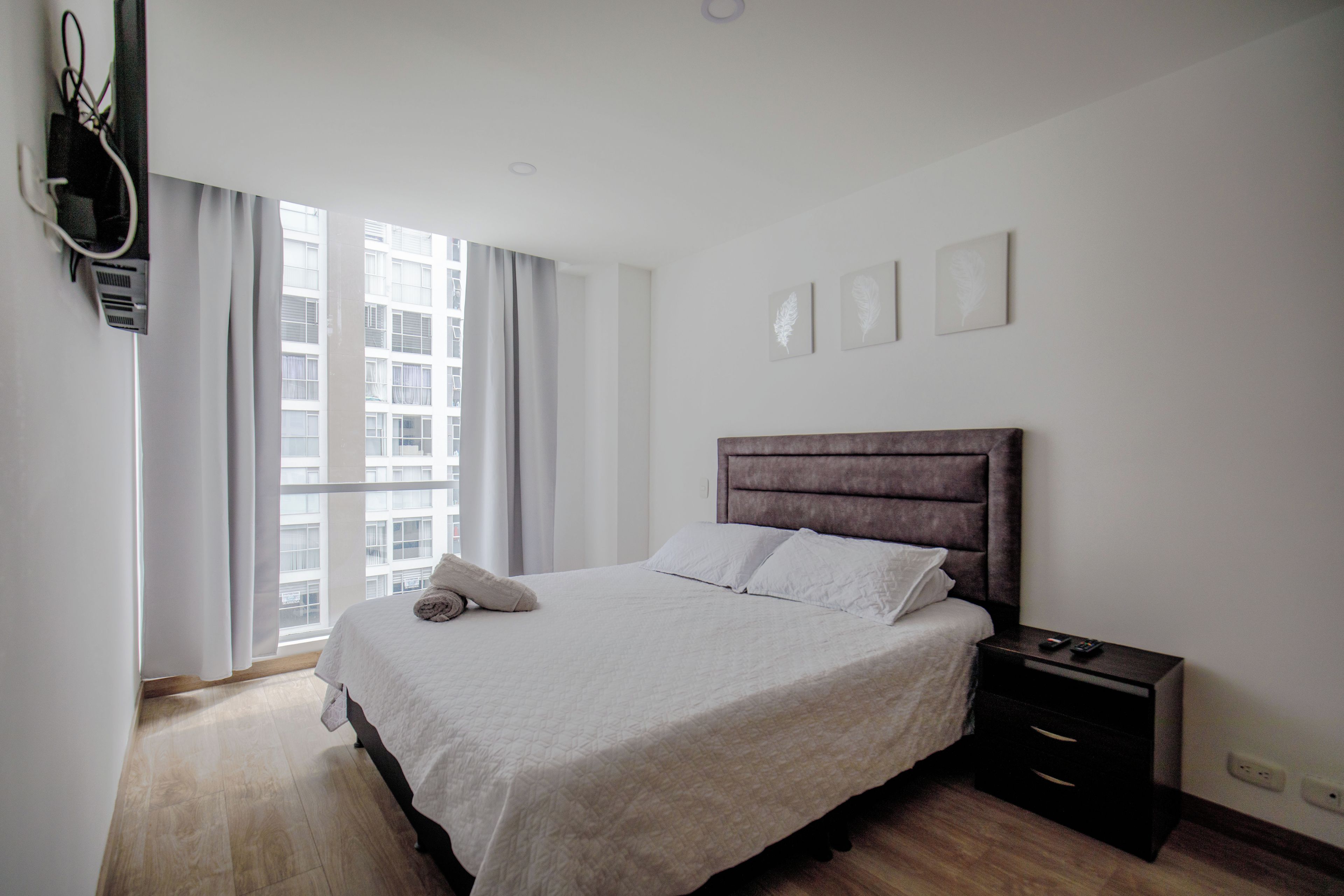 A bedroom with a bed, a nightstand, a window with curtains, and a wall-mounted TV.