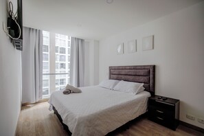 Un dormitorio con cama, mesita de noche, ventana con cortinas y televisor montado en la pared.