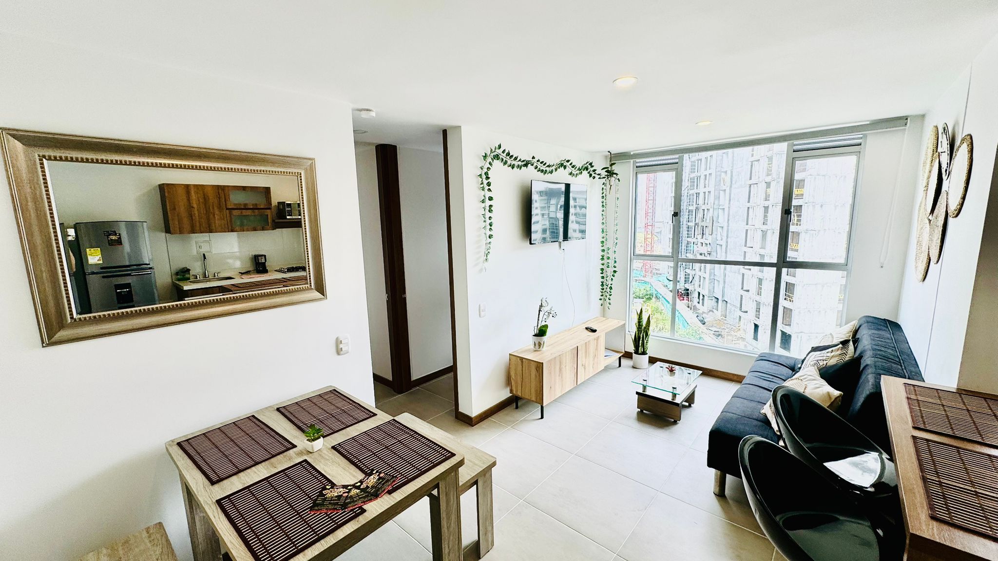 A modern living room with a large window, a sofa, a dining table, and a kitchen area visible through a mirror.
