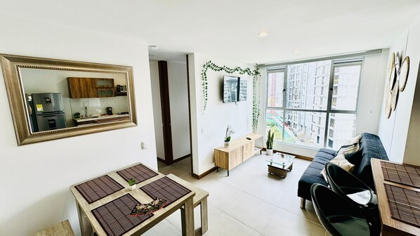 Una sala moderna con un ventanal grande, un sofá, una mesa de comedor y una cocina visible en un espejo.
