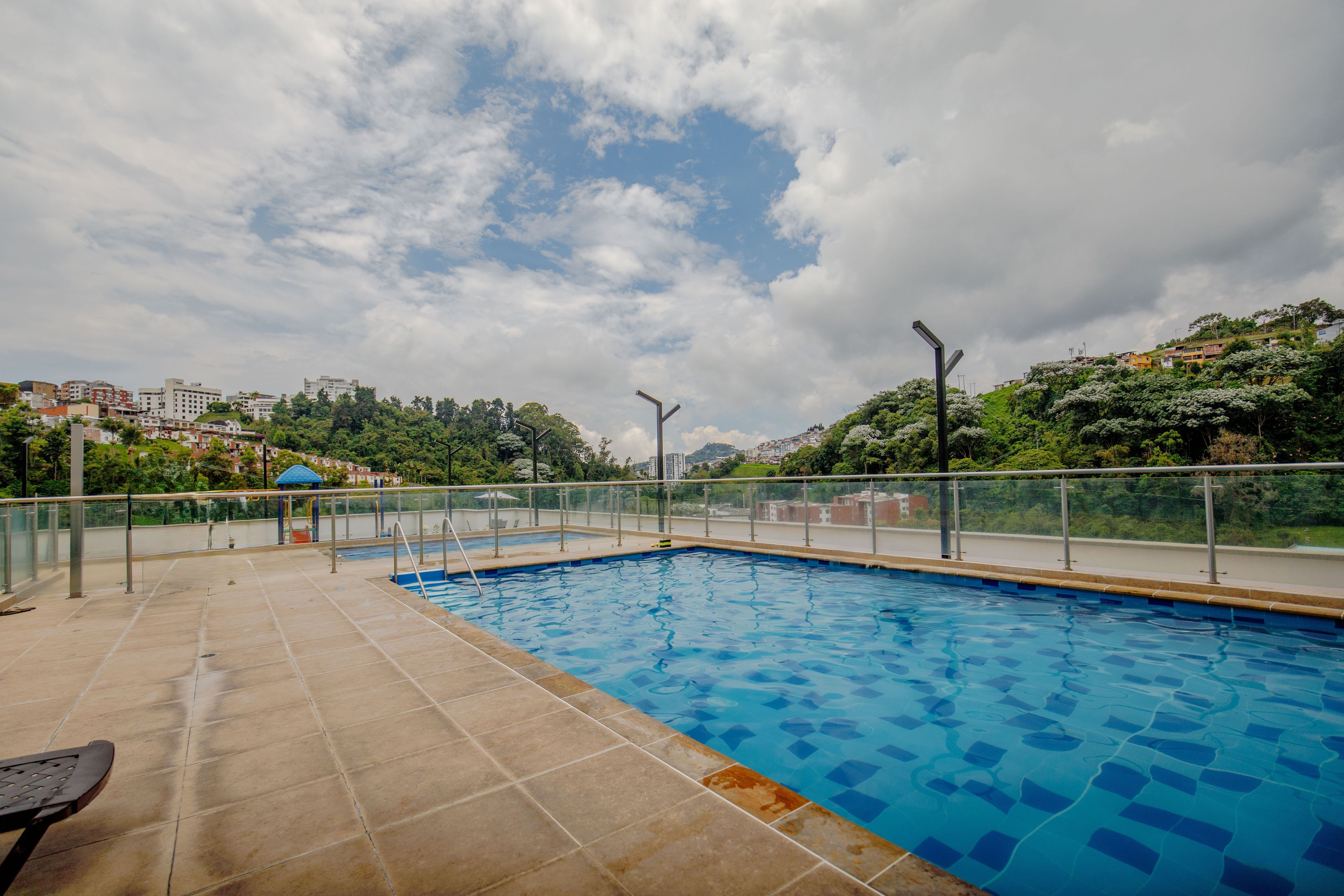 A swimming pool with a tiled deck, metal railings, and a view of a residential area with buildings and trees in the background.