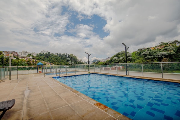 Una piscina con solárium empedrado, barandillas metálicas y vista a una zona residencial con edificios y árboles al fondo.