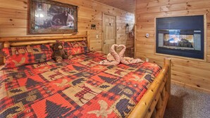 Een slaapkamer in een houten blokhut, met een bed, een teddybeer, een sprei, een open haard en een lijst met een foto aan de muur.