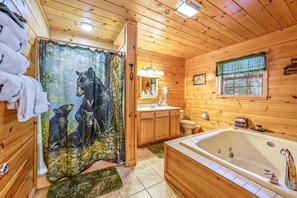 Une salle de bain rustique, située dans une cabane en bois, comprenant une baignoire, un lavabo, des toilettes et un rideau de douche orné d’un motif d’ours.