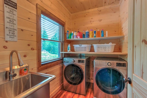 A compact laundry room with two washing machines, a sink, and a window with blinds. - A Rustic Escape-Theater Room/Handicap Ramp/Hot tub/2 fireplaces/2 king suites (Sevierville)