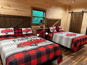 Quarto estilo cabana com duas camas, cada uma com manta xadrez preta e vermelha com desenho de carro, uma janela com persianas e teto de madeira.
