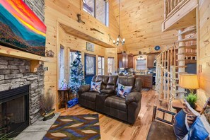 A cozy cabin interior with a fireplace, a leather sofa, a Christmas tree, and a kitchen area. - Duck Duck Goose-Riverfront cabin, Hot Tub overlooking river, fishing nearby (Pigeon Forge)