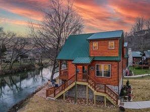 A log cabin with a green roof, a deck, and a staircase leading to a second level. - Duck Duck Goose-Riverfront cabin, Hot Tub overlooking river, fishing nearby (Pigeon Forge)