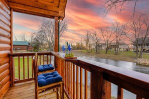 Un terrazza in legno con vista su un fiume e delle case, una sedia con cuscino e un tavolo con bicchieri e grappoli d'uva.