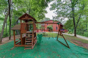 A wooden playground with a slide and swings, surrounded by trees and a red building in the background. - Dances with Wolves- Spacious Cabin, Hot tub, Swing, Large yard (Sevierville)