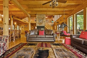 A spacious lodge interior with a central fireplace, wooden beams, a hanging chair, and a staircase leading to an upper level. - Dances with Wolves- Spacious Cabin, Hot tub, Swing, Large yard (Sevierville)