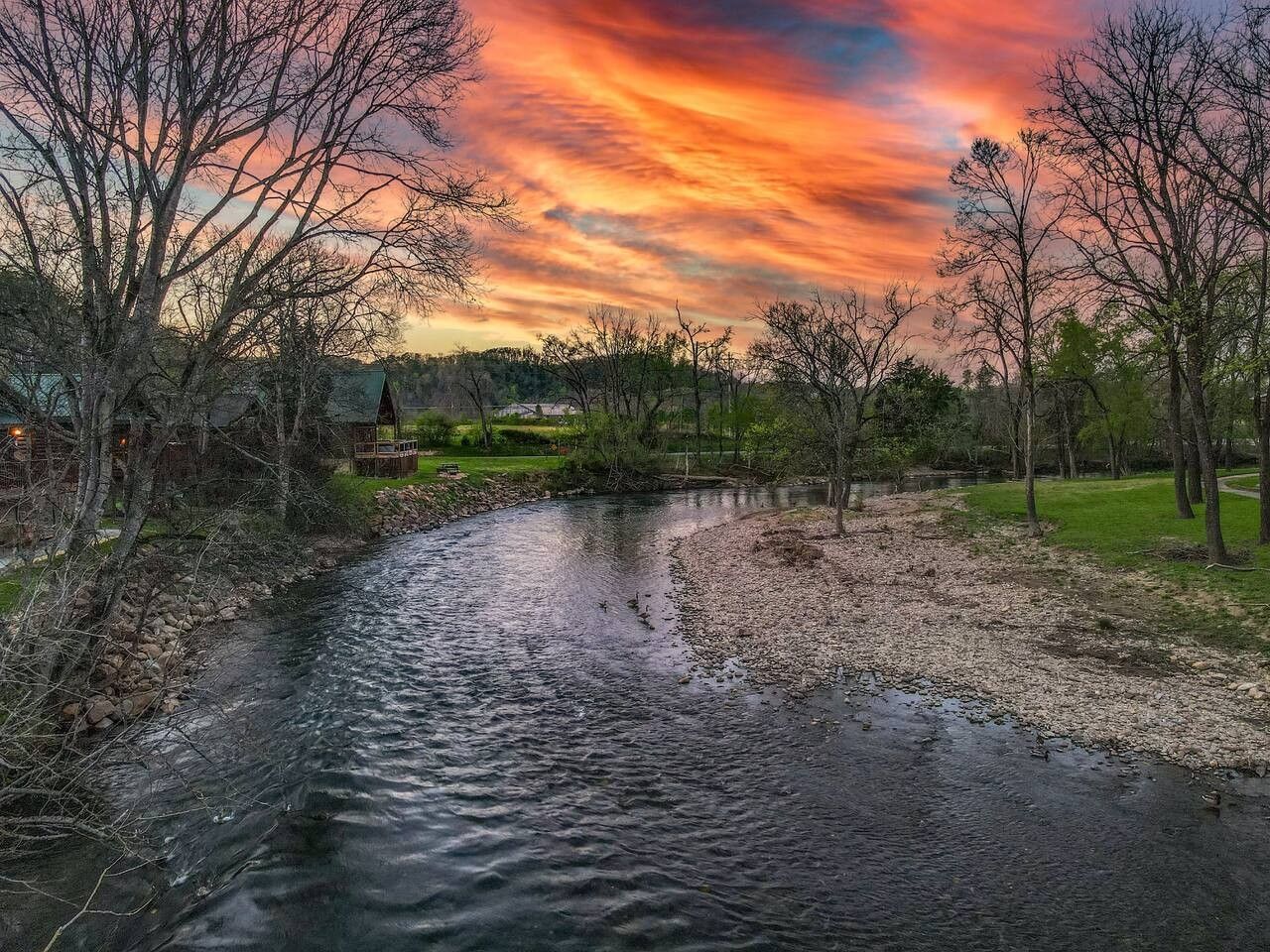 Ein Fluss mit Bäumen an beiden Ufern, eine Wiese und ein Gebäude im Hintergrund.