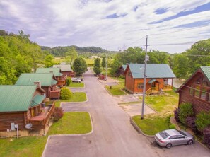 A row of log cabins with green roofs, a paved driveway, and a car parked on the right. - A Barrel of Fun-riverfront cabin, fishing nearby (Pigeon Forge)