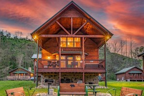 A wooden cabin with a balcony, stone foundation, and a grassy yard with benches. - A Barrel of Fun-riverfront cabin, fishing nearby (Pigeon Forge)