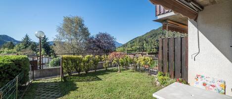 Un espace patio aménagé avec une table et des chaises, entouré d’un jardin où poussent différentes plantes, et offrant une vue sur une montagne.