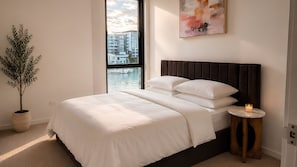 A bedroom with a bed, a bedside table with a candle, a potted plant, and a window with a view of buildings and water. - Marina Prestige, Hope Island (Hope Island)