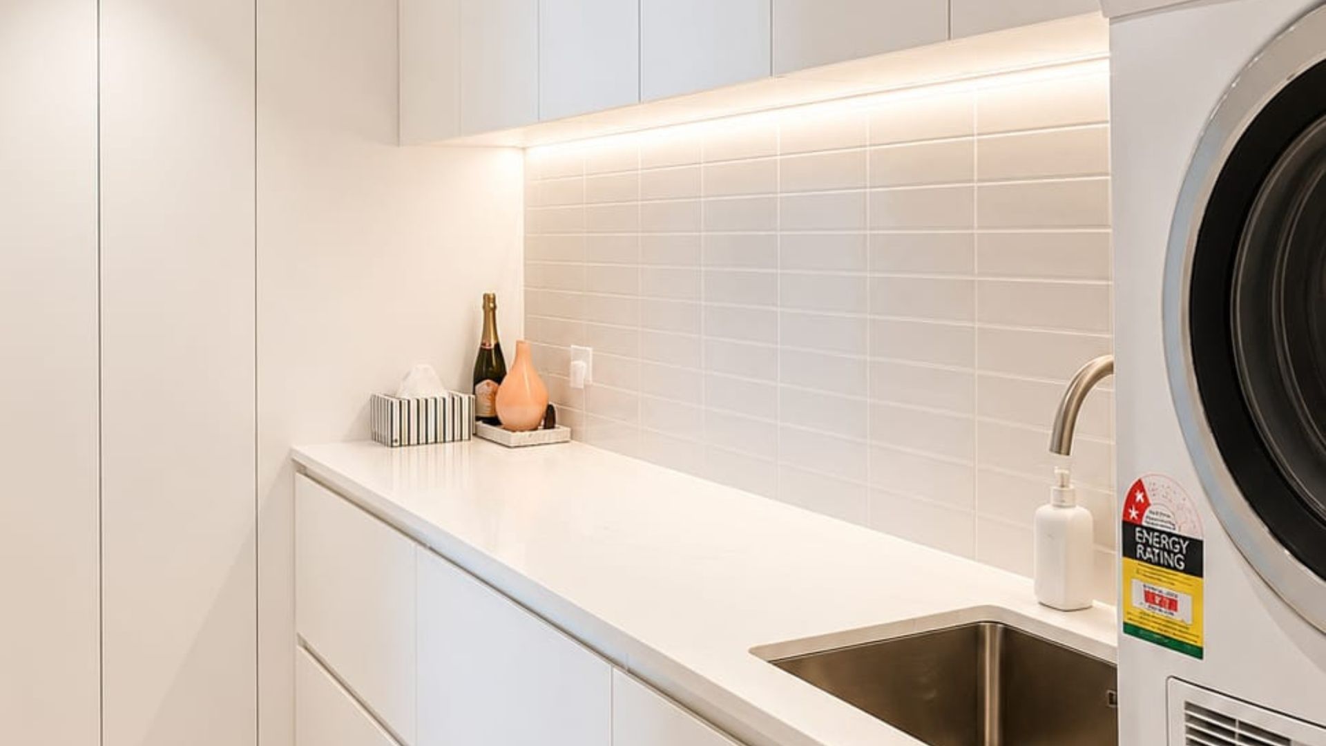 Eine moderne Küche mit integriertem Waschmaschine, Spüle und einer Arbeitsplatte, auf der eine Champagnerflasche und eine Taschentuchbox stehen.