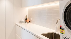 A modern kitchen with a built-in washing machine, a sink, and a countertop with a bottle of champagne and a tissue box. - Marina Prestige, Hope Island (Hope Island)