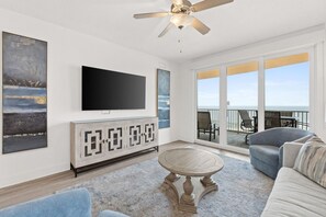 A living room with a flat-screen TV, a ceiling fan, a large window with a view, and a balcony with outdoor seating. - Walk to the Beach • Resort Pools • Pier Park (Panama City Beach)