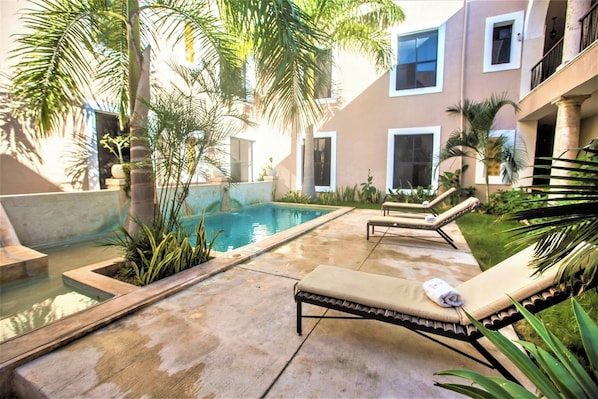 A pool area with lounge chairs and a small pool, surrounded by palm trees and a building with large windows. - Colonial Mérida Outside, Poolside Relaxation Inside | 2 Beds (Mérida)
