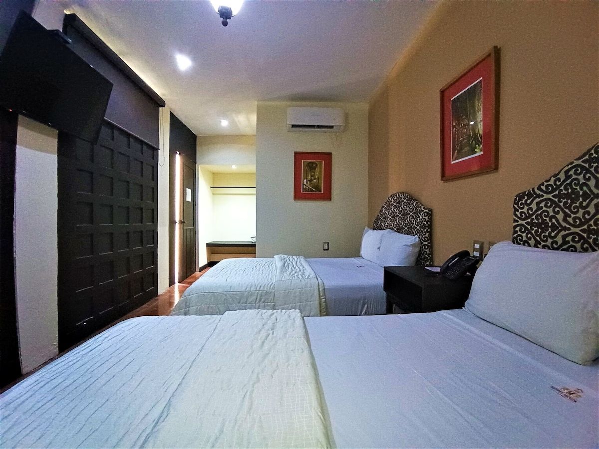 Quarto de hotel com duas camas, uma mesinha de cabeceira, ar condicionado e uma porta com painel de segurança.