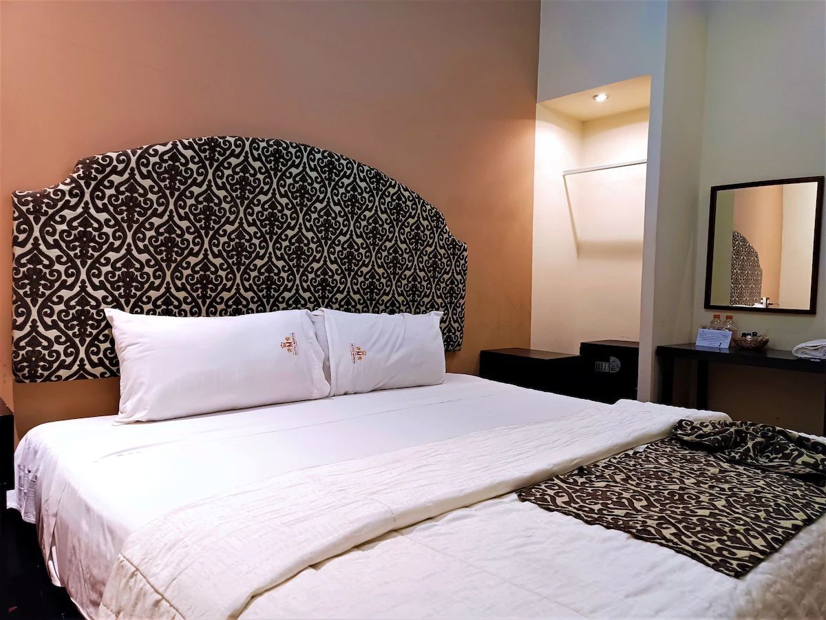 Quarto de hotel com uma cama grande, cabeceira estampada, mesa de cabeceira com abajur e um espelho na parede.