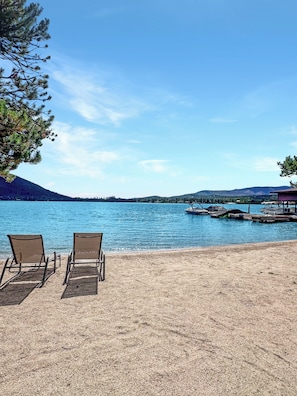 Uma praia com duas cadeiras de praia viradas para um lago calmo, cercada por árvores e um píer com barcos.